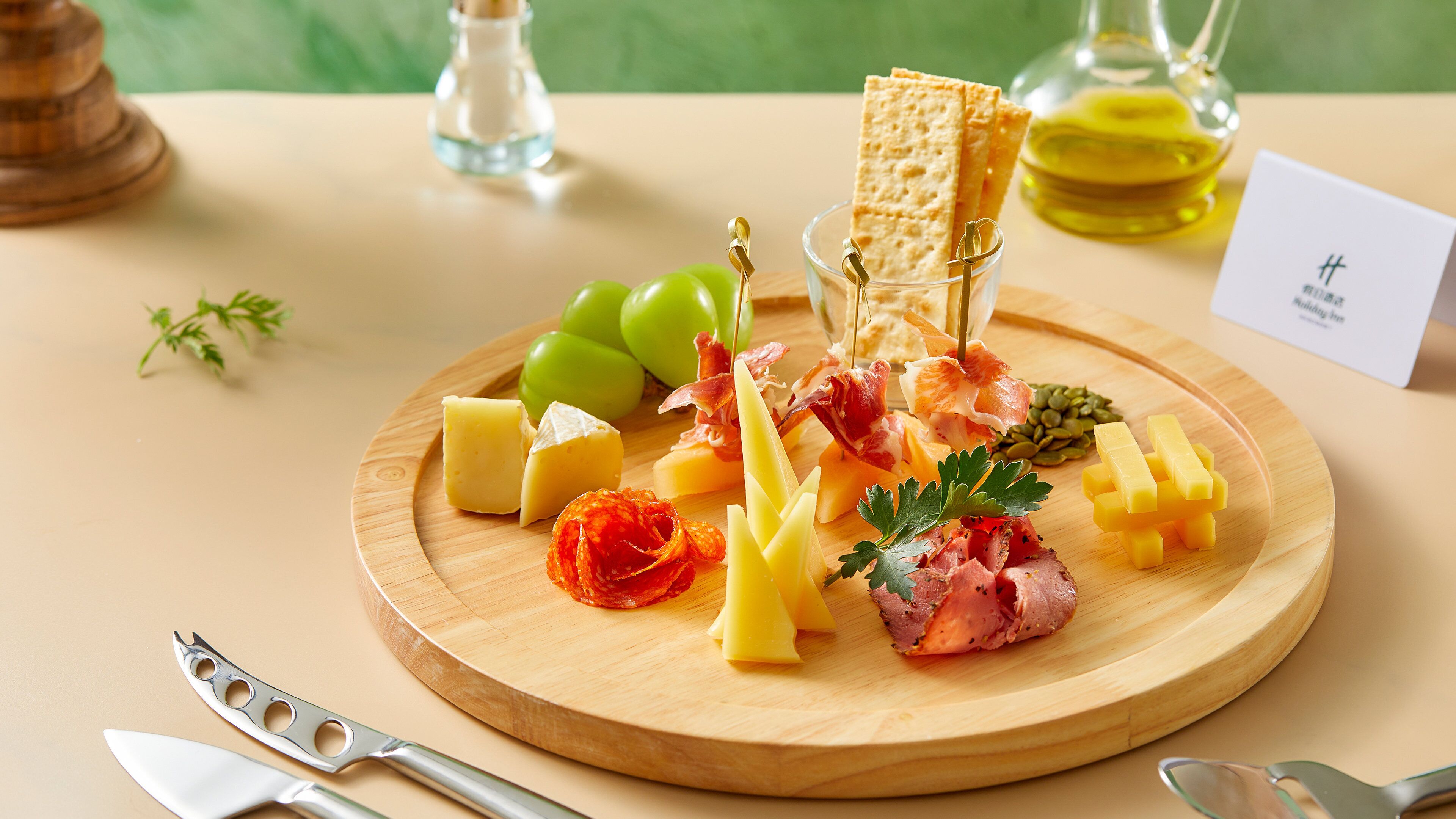 A wooden board with various cheeses, cured meats, and crackers, accompanied by a bottle of oil and a card.