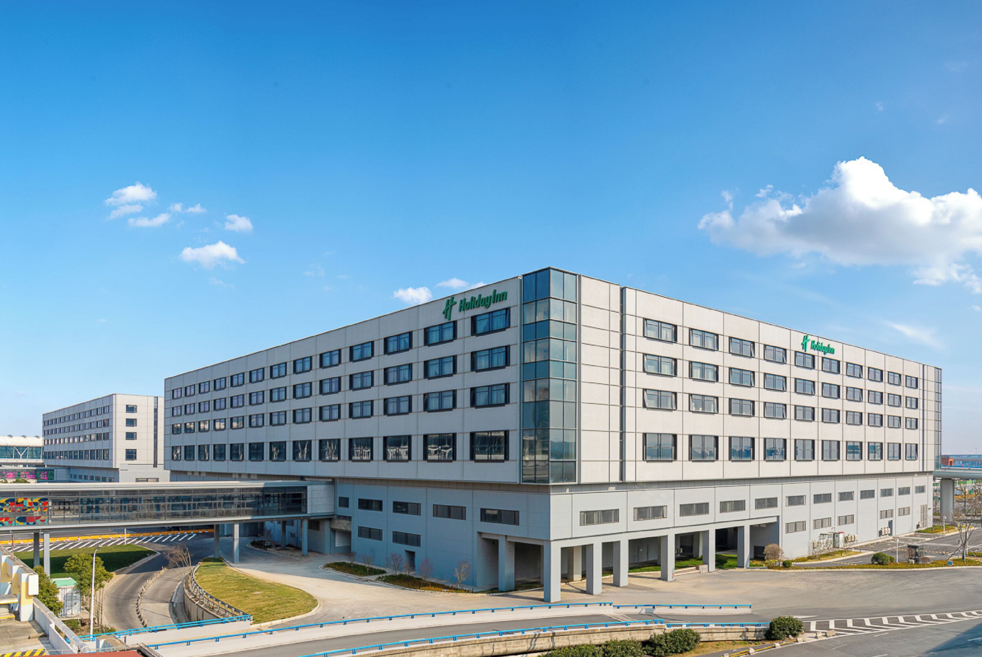 Un hotel Holiday Inn moderno con techo plano, amplias ventanas y un estacionamiento con barrera azul.