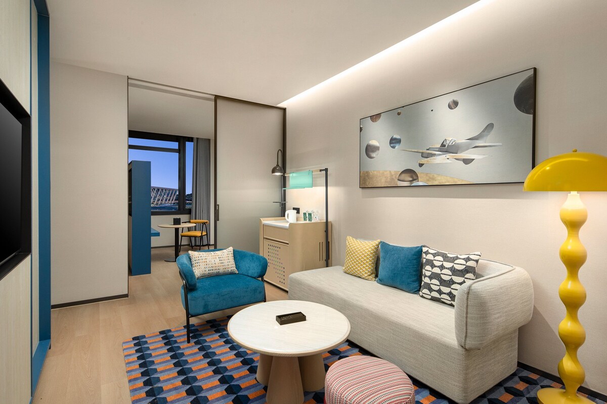 A modern hotel room with a sofa, armchair, round table, and a TV.