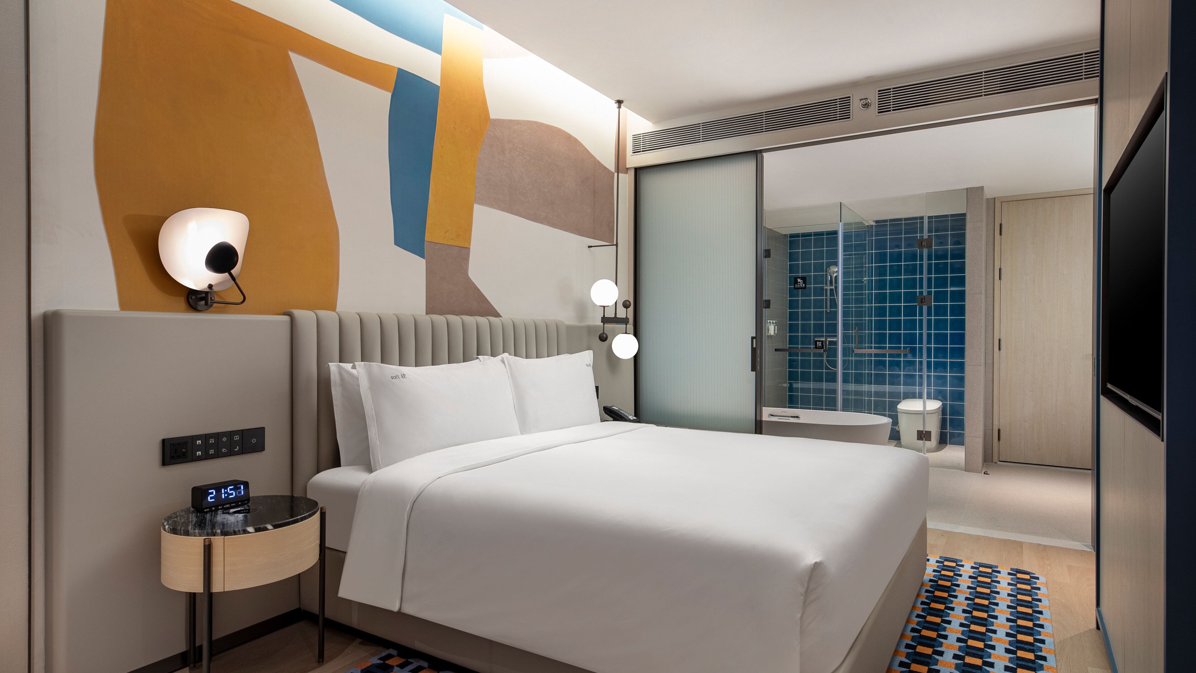 A modern hotel room with a large bed, bedside controls, a digital clock, a small table with a lamp, and a bathroom with a glass shower enclosure.