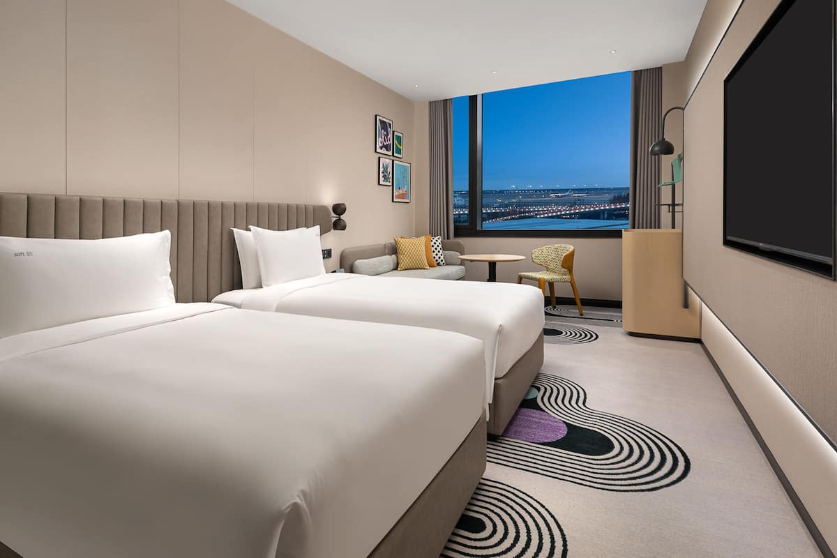 A hotel room with two beds, a sofa, a small table, a chair, and a large window with a city view.