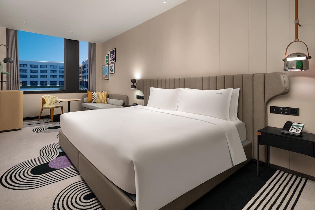 A modern hotel room with a large bed, a desk, a chair, and a view of the cityscape.