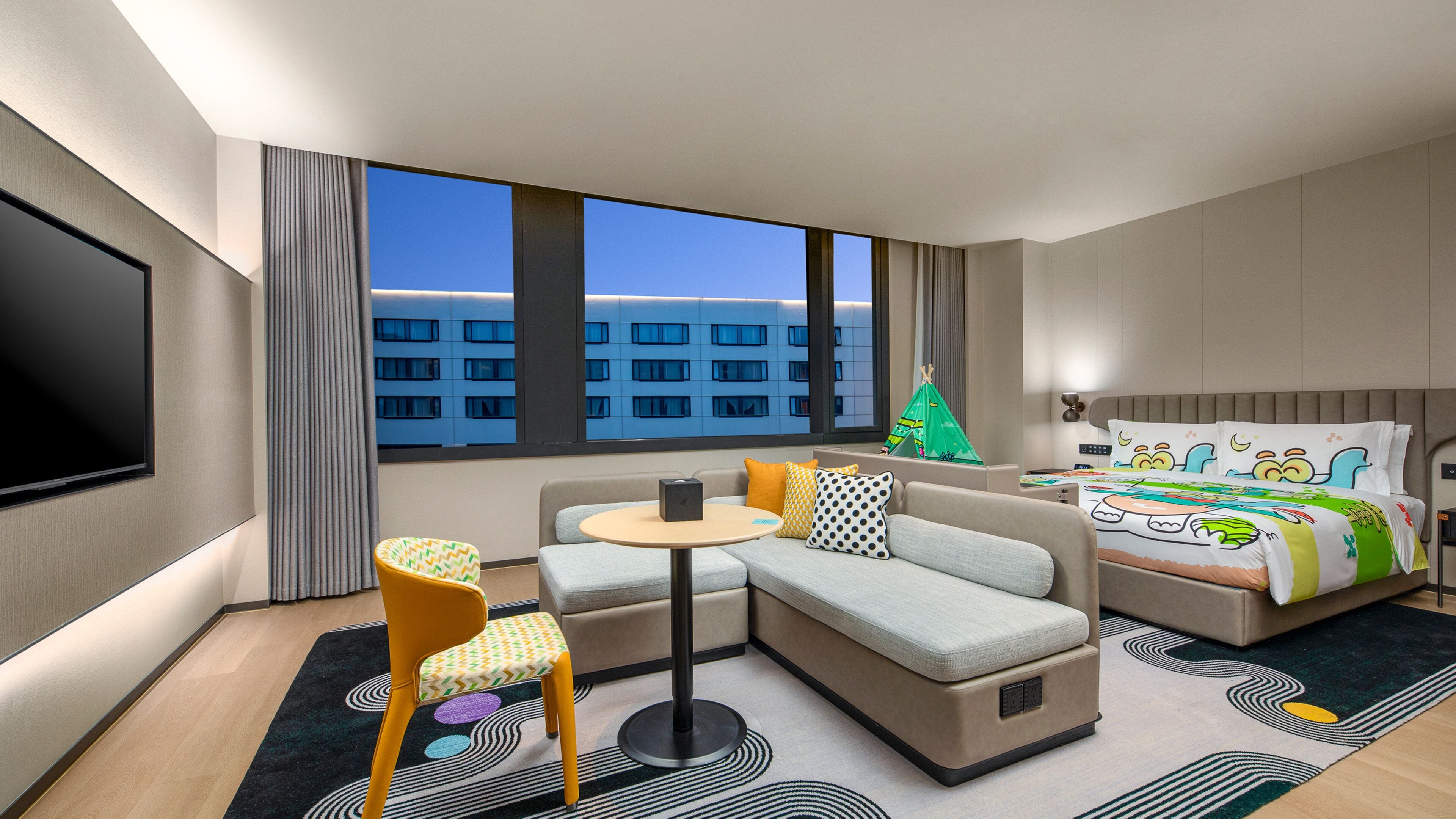 A modern hotel room with a sofa, a small table, a chair, a bed with colorful bedding, a TV, and a view of a building through large windows.