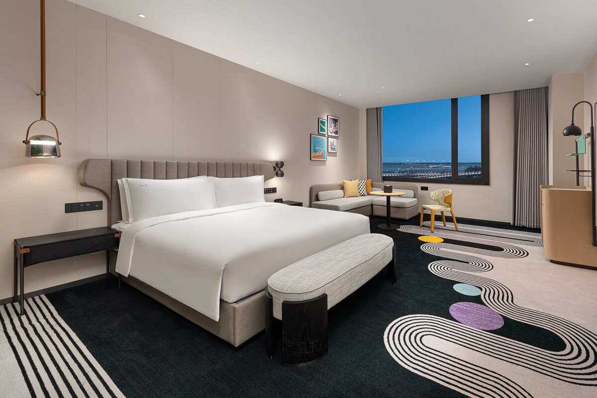 A modern hotel room with a large bed, a sofa, a desk, and a view of the cityscape.