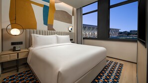 Una habitación de hotel moderna con una cama grande, mesitas de noche, una lámpara y vistas de la ciudad a través de amplios ventanales.