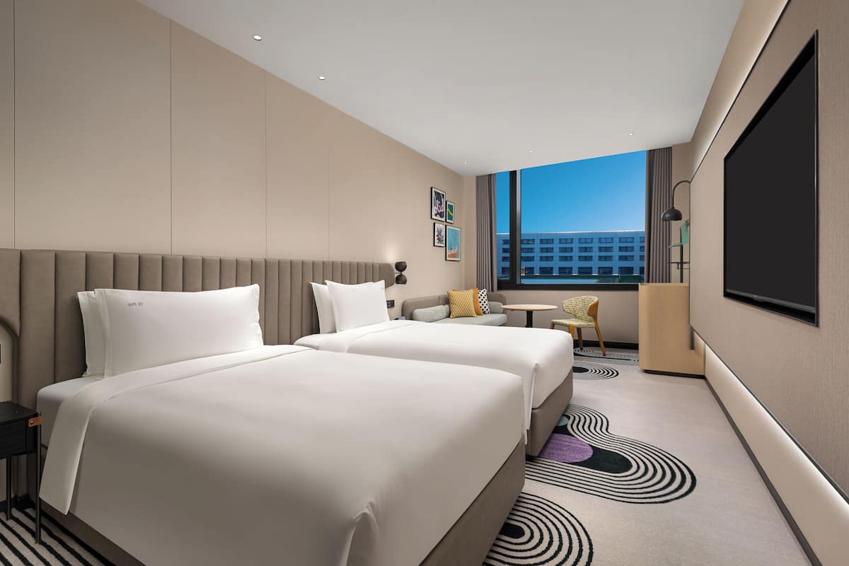 A hotel room with two beds, a small table, a chair, a TV, and a view of the city.
