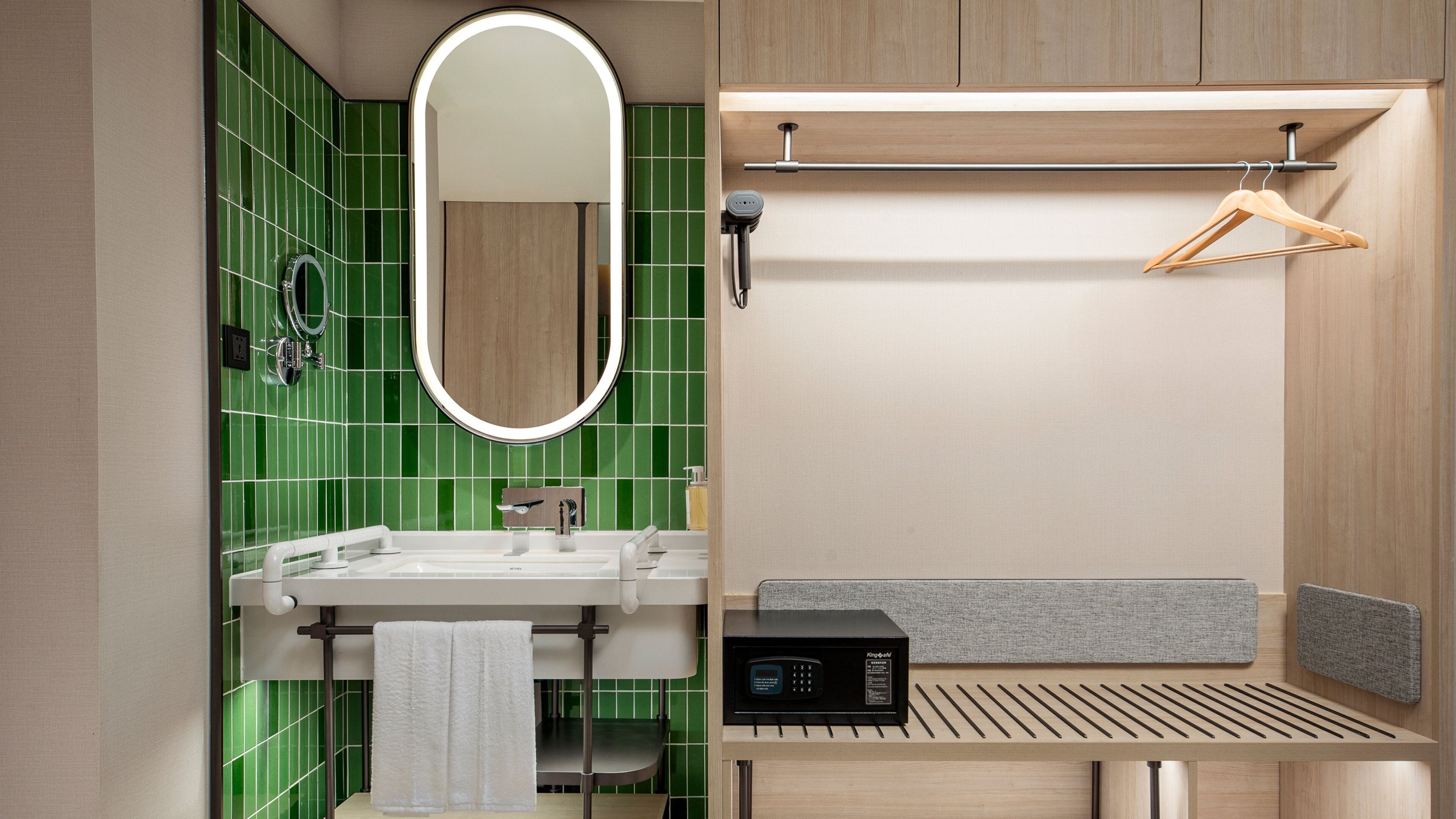 A modern bathroom with a green tiled wall, a round mirror, a sink with a towel, and a shower area with a wooden bench and a towel rack.