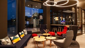 A modern lounge area with a sofa, armchairs, and round tables, featuring a city view through large windows. - Holiday Inn Express & Suites Monheim Am Rhein by IHG (Monheim)