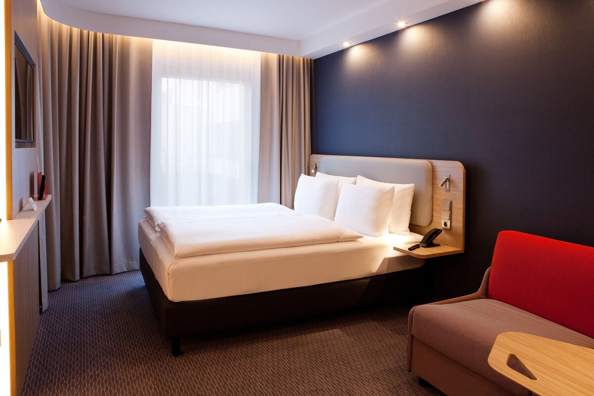 A modern hotel room with a large bed, a bedside table with a lamp, a red sofa, and a large window with curtains.
