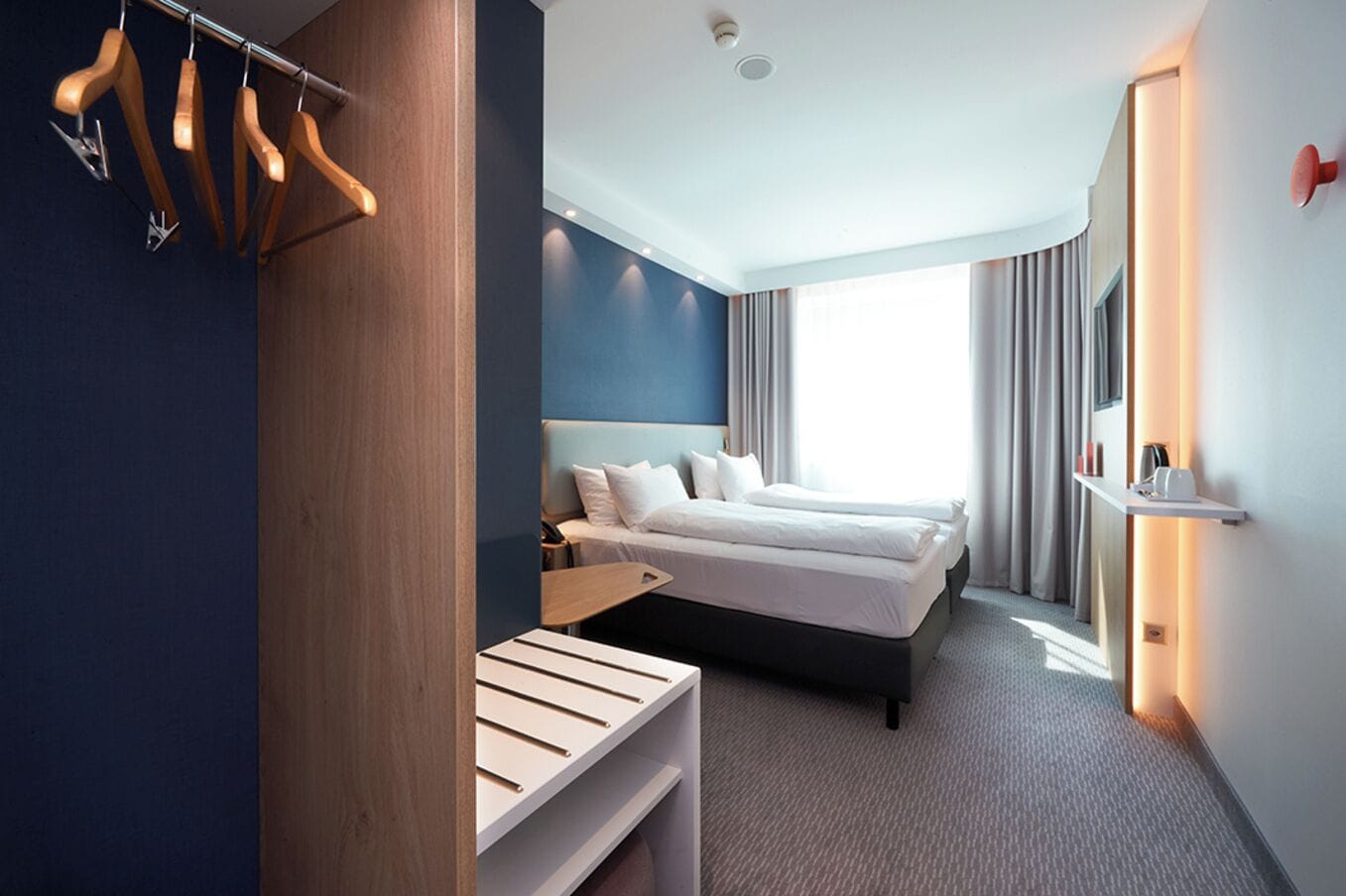 A modern hotel room with a bed, bedside tables, a desk, and a wardrobe with wooden hangers.
