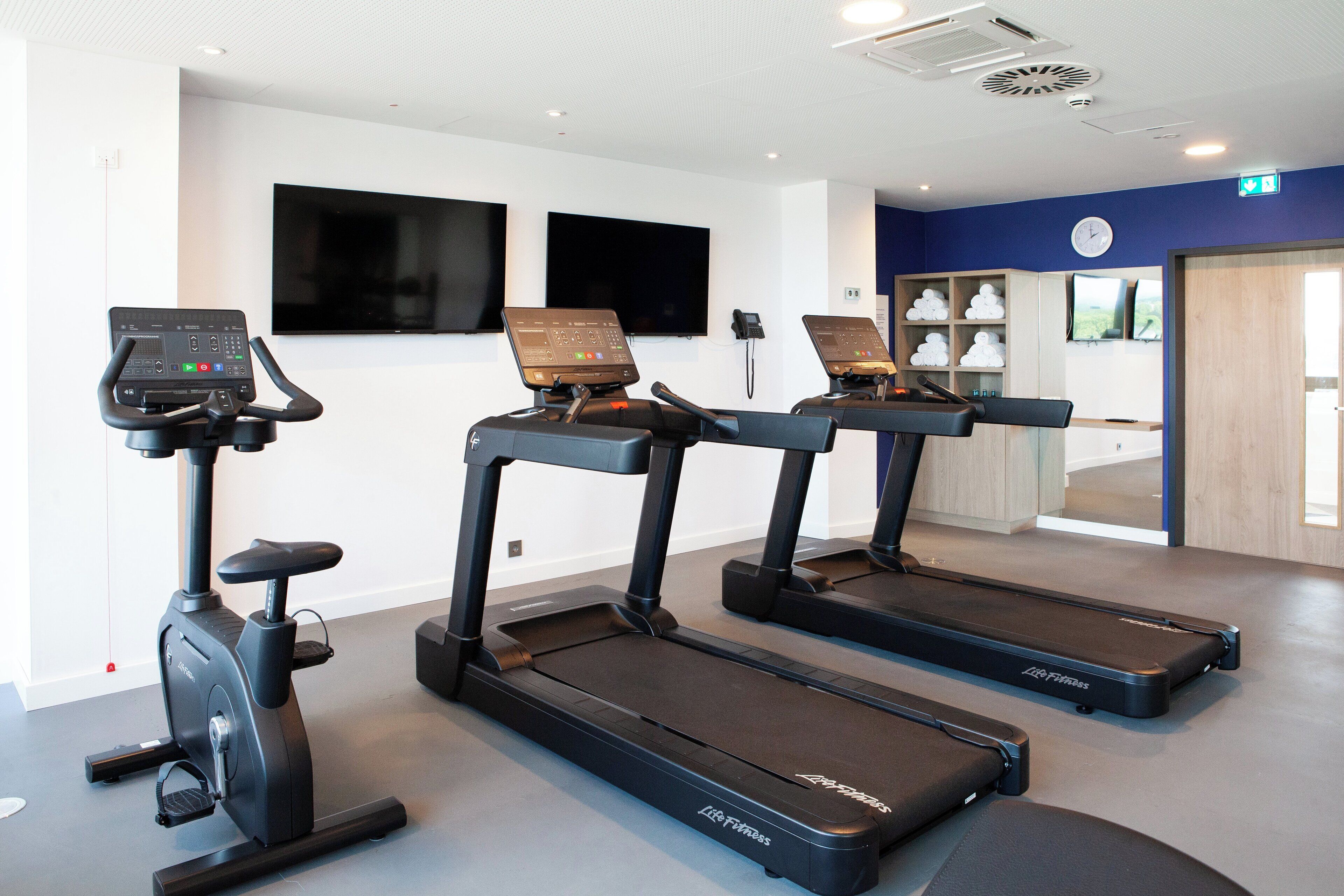 Un gimnasio con cintas de correr y una máquina elíptica, un televisor en la pared y un reloj.