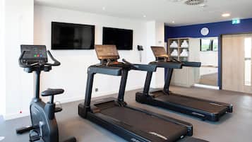 Un gimnasio con cintas de correr y una máquina elíptica, un televisor en la pared y un reloj.