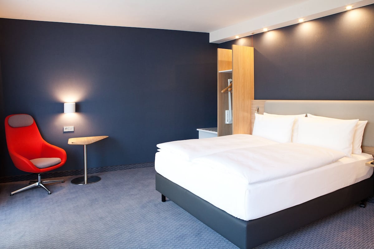 A modern hotel room with a large bed, a red chair, a small table, and a wall-mounted light.