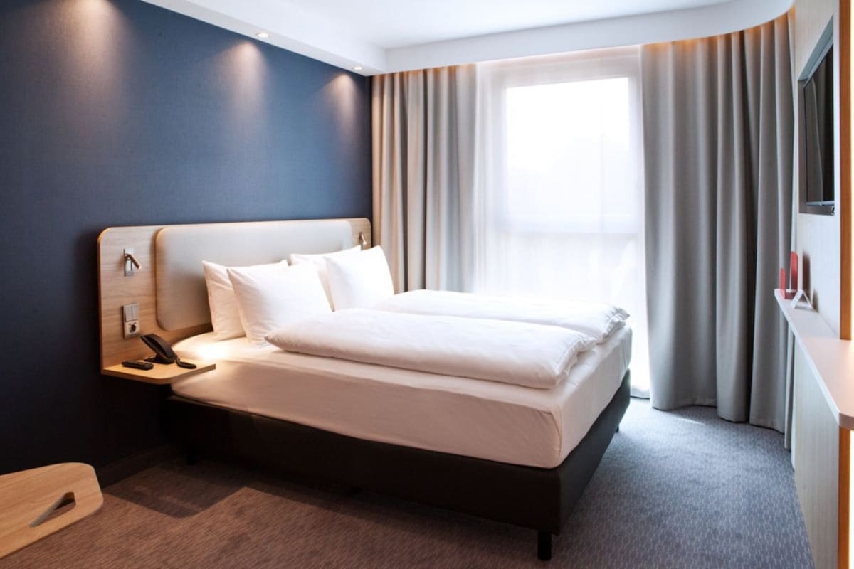 A modern hotel room with a large bed, a bedside table with a phone, and a window with curtains.