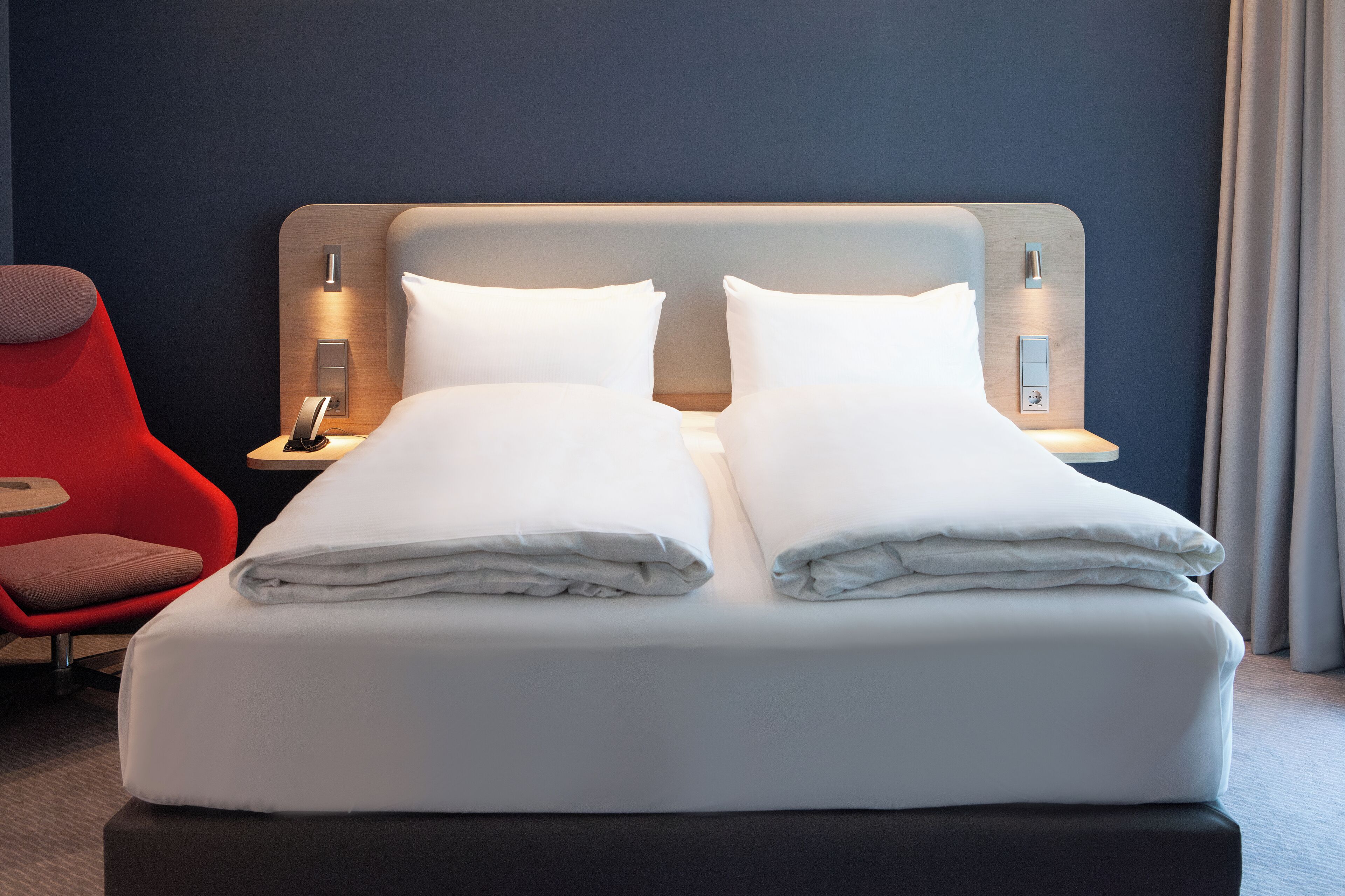 Una cama doble con ropa blanca impecable, una silla roja y una mesita de noche con un teléfono y una lámpara.