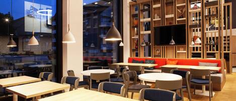 A modern cafe with round tables and chairs, a wooden bookshelf, and a TV screen.