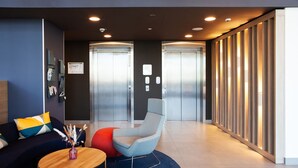 A modern lobby with a blue sofa, a round wooden table, and a grey armchair. There are two elevators and a wooden partition with vertical slats. - Holiday Inn Express & Suites Monheim Am Rhein by IHG (Monheim)