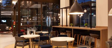 Un café moderno con mesas y sillas redondas, una barra de madera y ventanas amplias.