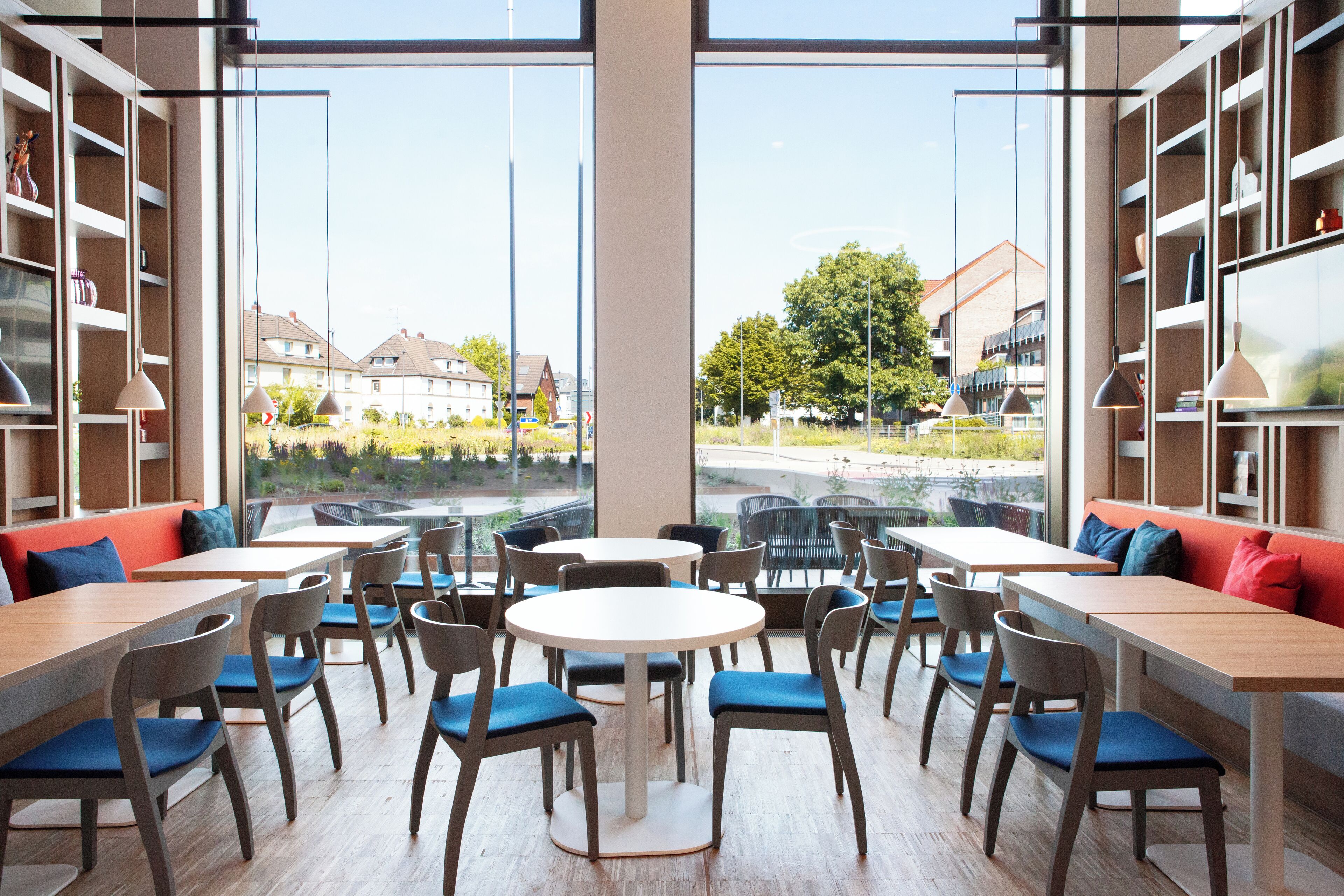 A modern cafe with round tables and chairs, large windows offering a view of the street and buildings, and shelves with various items.
