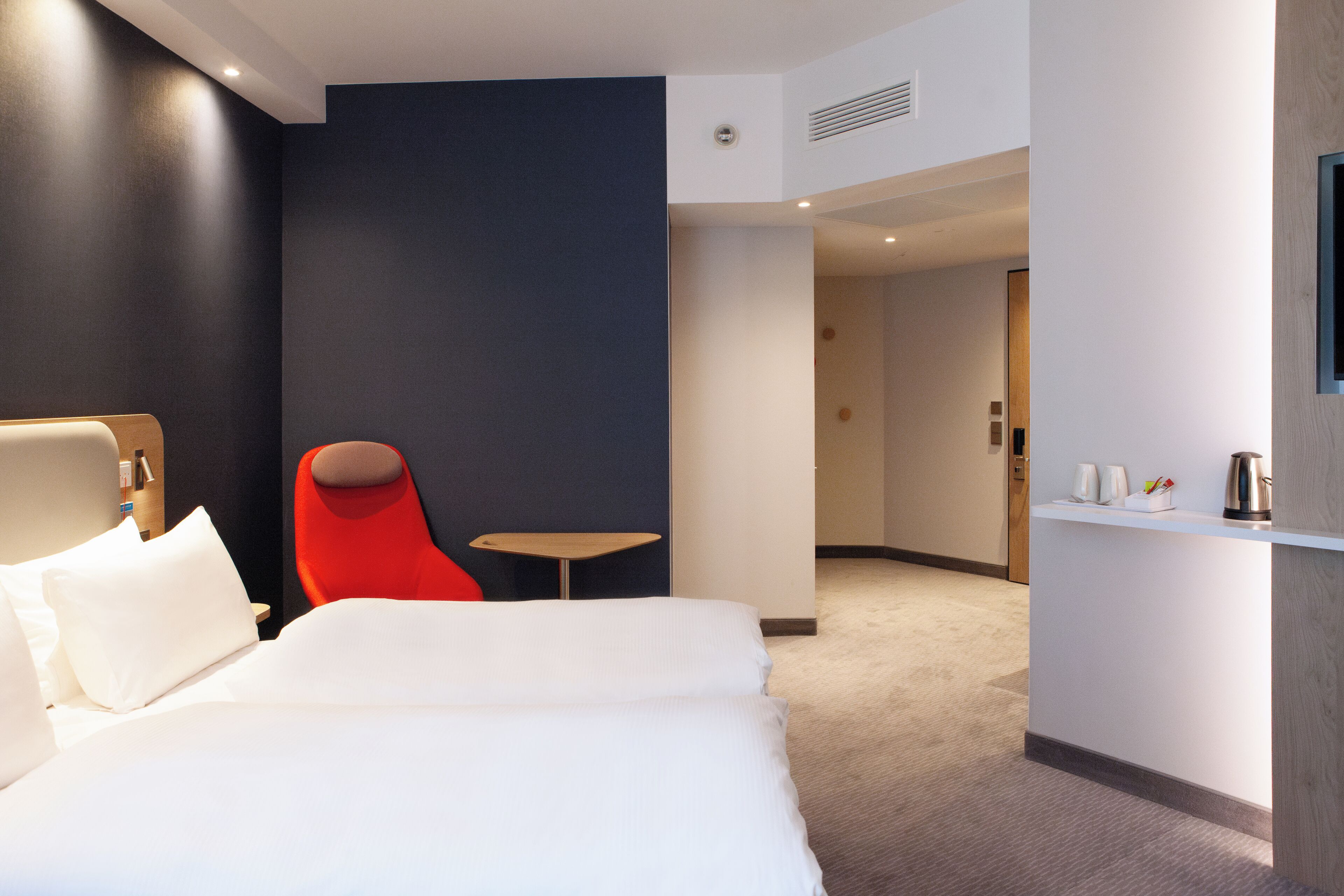 Ein Hotelzimmer mit einem großen Bett, einem roten Sessel, einem kleinen Tisch und einer Küchenzeile mit einer Kaffeemaschine.