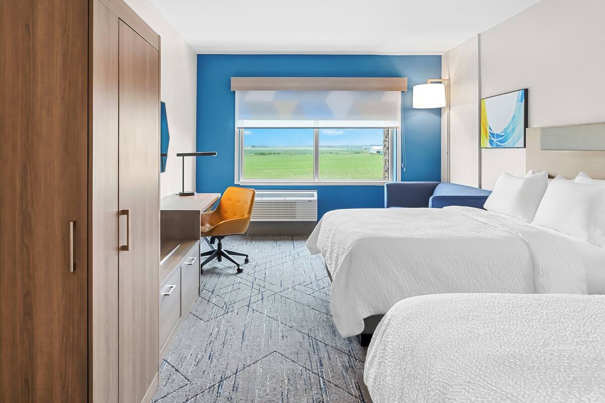 Ein Hotelzimmer mit zwei Betten, einem Schreibtisch, einem Stuhl und einem großen Fenster mit Blick auf ein grünes Feld.