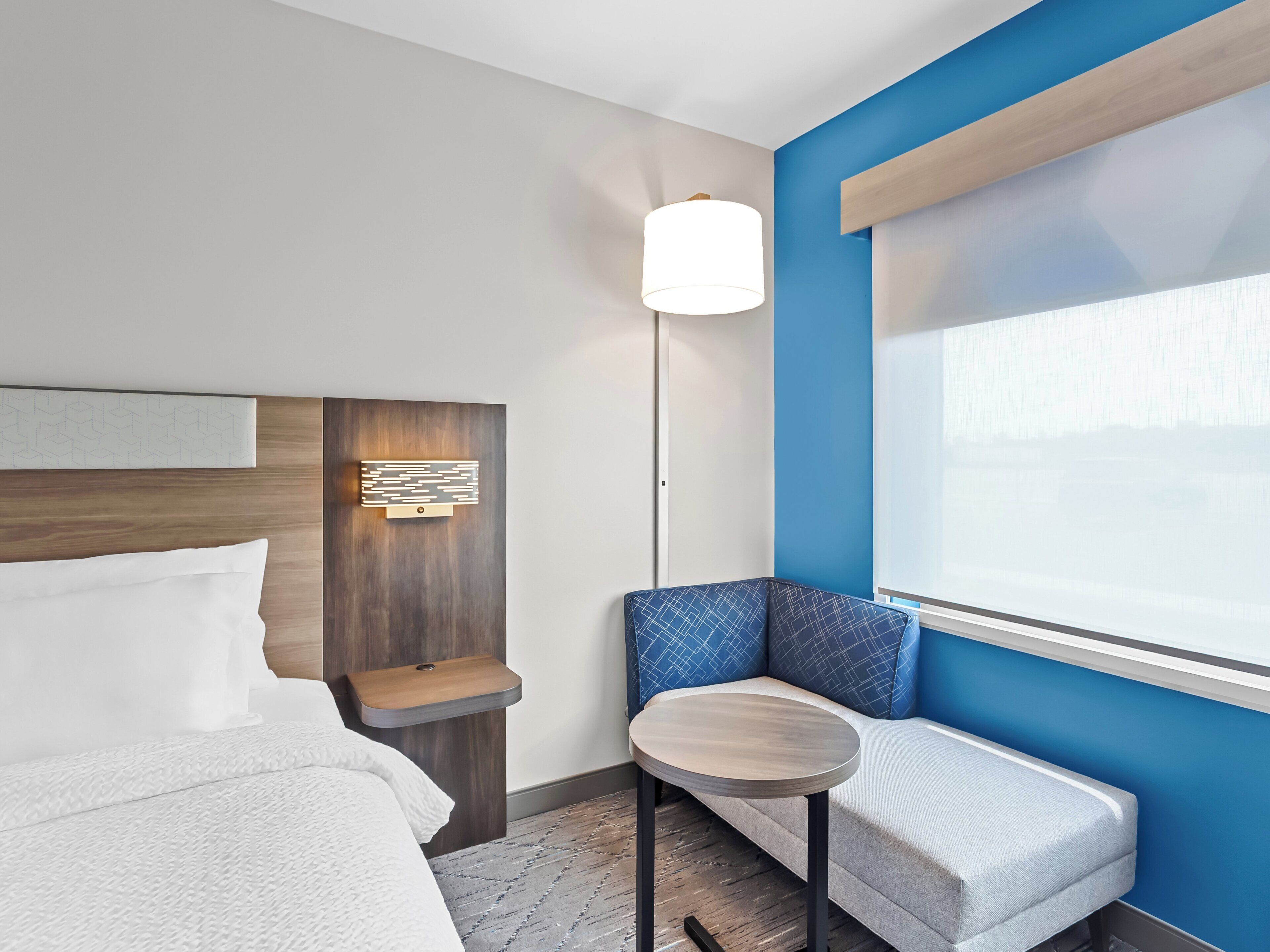 Une chambre d’hôtel avec un lit, une petite table ronde, un banc et une grande fenêtre.