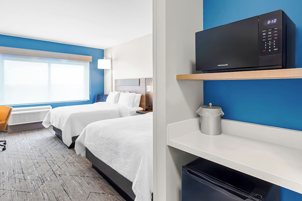 Hotellihuone, jossa on kaksi sänkyä, mikroaaltouuni, roskakori ja ikkuna, jossa on sälekaihtimet.