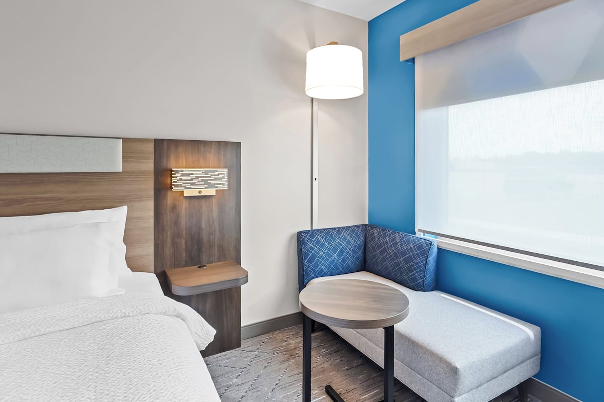 Hotellihuone, jossa on sänky, pieni pyöreä pöytä, penkki ja suuri ikkuna.