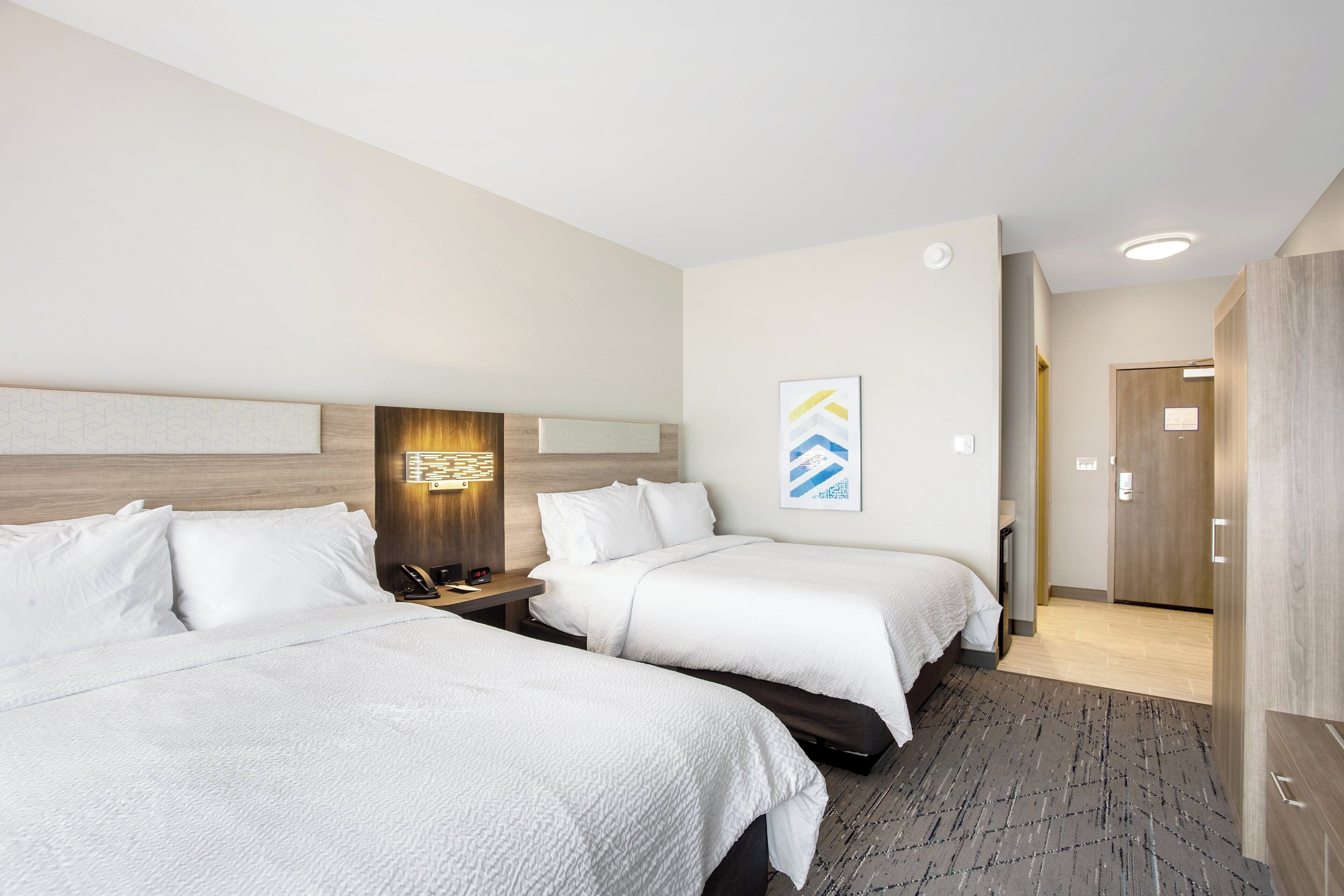 Habitación de hotel con dos camas, cabecero de madera, mesita de noche, lámpara de pared y una puerta que da a otra habitación.