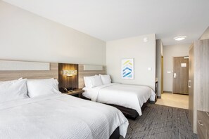 A hotel room with two beds, a wooden headboard, a nightstand, a wall-mounted lamp, and a door leading to another room.