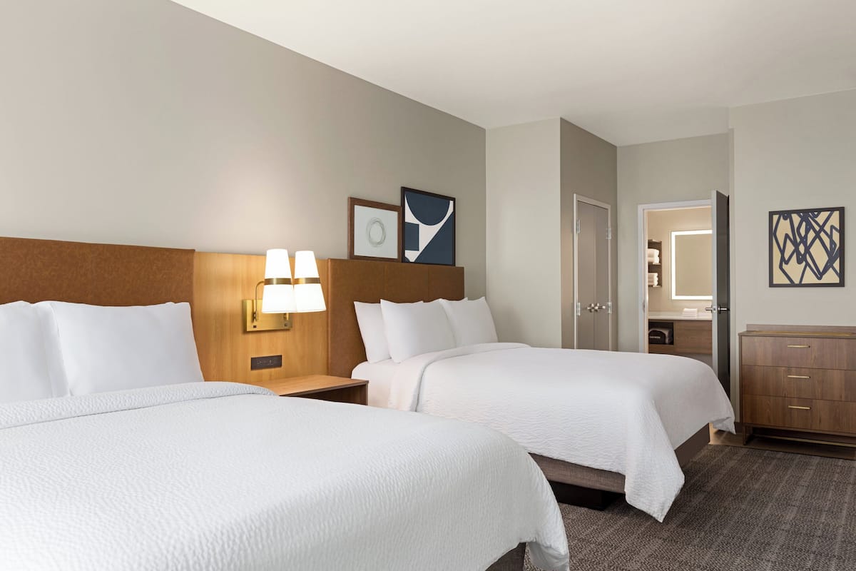 Une chambre d’hôtel avec deux lits, une tête de lit en bois, une table de chevet avec une lampe, une commode et une porte donnant sur une salle de bain.