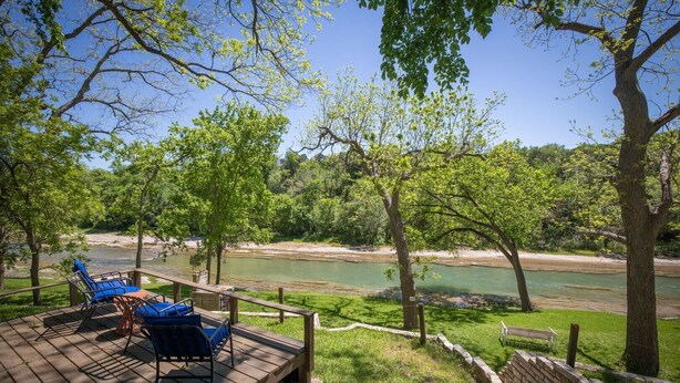 A wooden deck with outdoor seating, overlooking a river and surrounded by trees. - PRIVATE and serene riverfront retreat - LARGE spacious yard with a cozy home!  (Canyon Lake)