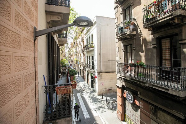 A narrow urban street with buildings on both sides, featuring balconies with potted plants, a pedestrian walkway, and a street lamp.