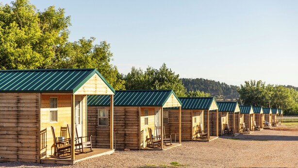 A row of log cabins with green roofs and wooden porches, each with a chair and a table. - Black Hills Station  (Piedmont)