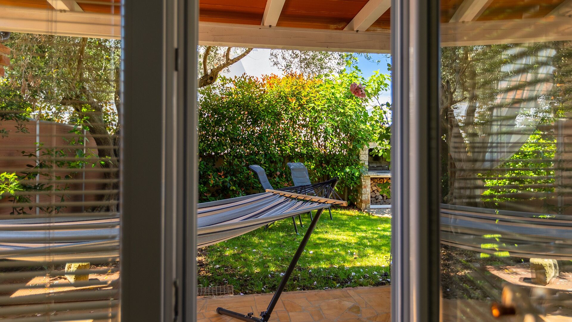 Un hamac sous une porte vitrée, une terrasse en pierre et un jardin agrémenté d’une cheminée en pierre.