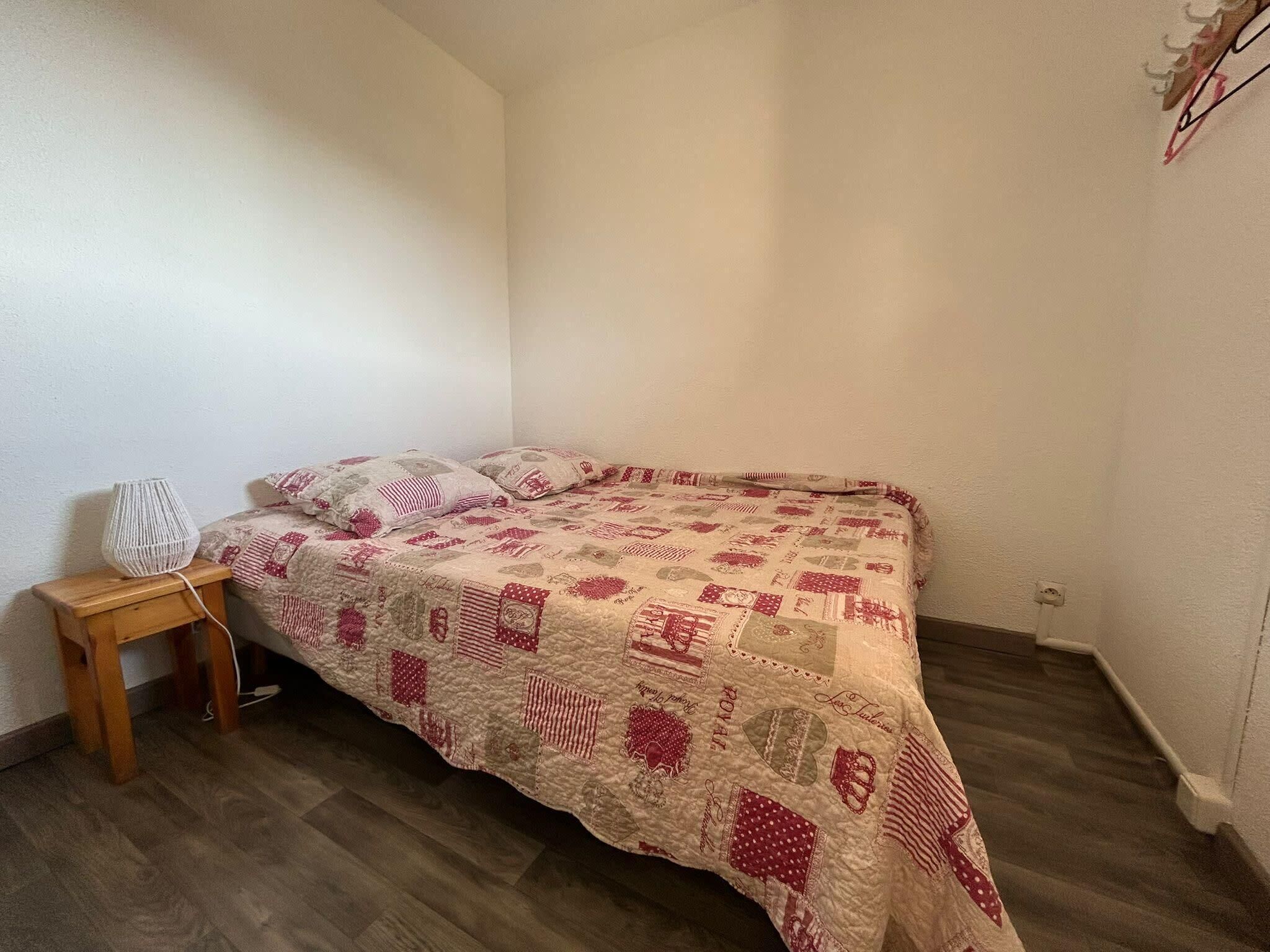 A small, neatly made bed with a quilted blanket featuring a patchwork design, a wooden nightstand with a lamp, and a plain white wall.