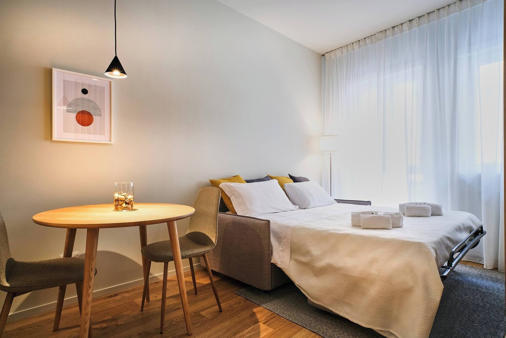 Une chambre d’hôtel avec un lit, une table de chevet, une chaise, une lampe et une fenêtre avec des rideaux.