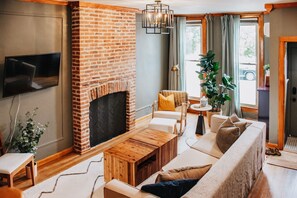 A living room with a brick fireplace, a wooden coffee table, a sofa, and a TV mounted on the wall. - Baltimore's Luxury Rowhome Stay (Baltimore)