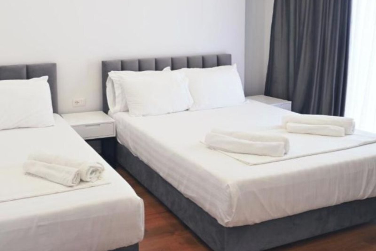 A hotel room with two beds, white linens, grey headboards, and a large window with dark curtains.