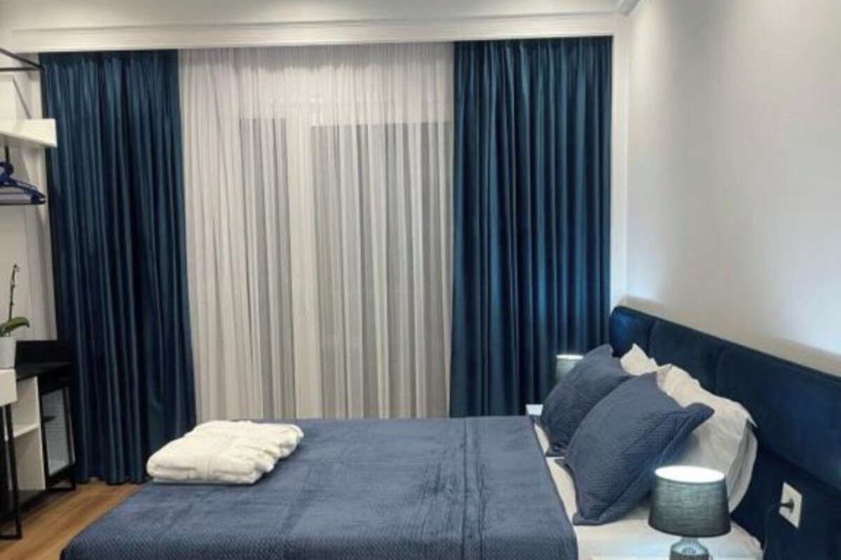 A modern bedroom with a large bed, dark blue curtains, a bedside table with a lamp, and a small plant.
