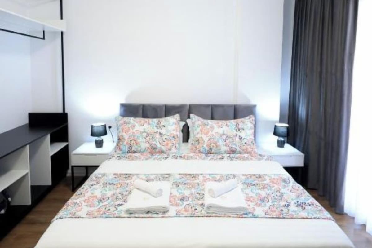 A modern hotel room with a neatly made bed, a bedside table with a lamp, and a large window with curtains.