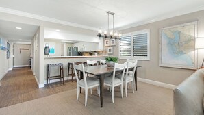 A dining area with a wooden table and white chairs, a hanging light fixture, and a map on the wall. - Sanibel Beachfront Luxury Condo Full Gulf View! 3BR, 2 BA! Covered Prkg! Sports! (Sanibel)