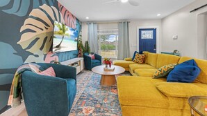 A living room with a tropical mural, a blue sofa, a yellow sofa, and a coffee table. - PalmParadise: 5BR Family&Pet Fun, HeatedPool&Games (West Palm Beach)