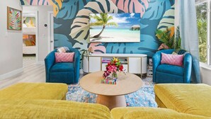 A modern living room with a tropical-themed wall mural, two blue armchairs, a round coffee table with a floral centerpiece, and a view of a beach through a large window. - PalmParadise: 5BR Family&Pet Fun, HeatedPool&Games (West Palm Beach)