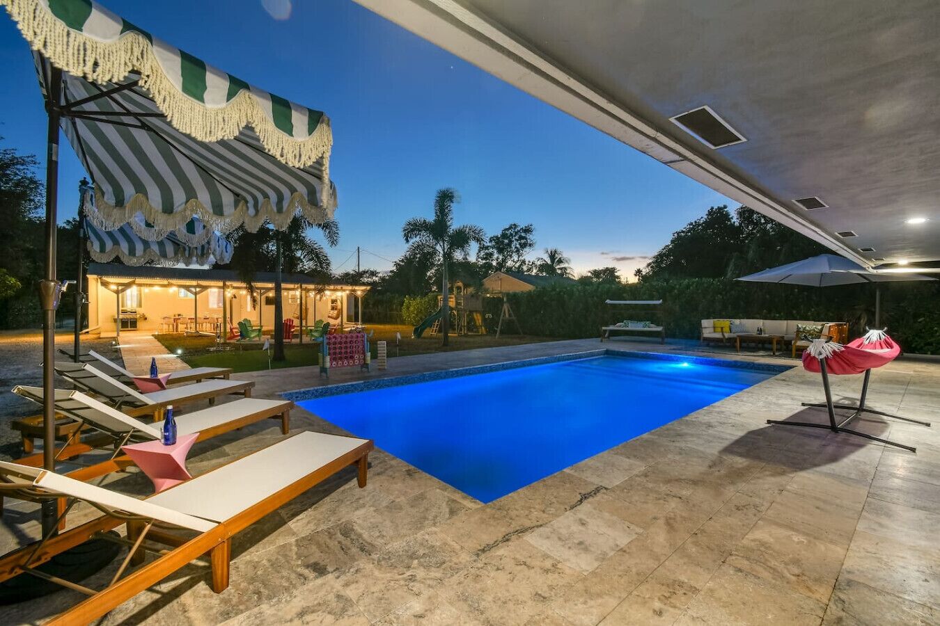 A swimming pool with lounge chairs and umbrellas, surrounded by a patio area with a covered seating area and a slide.