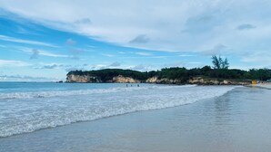 Ein Strand mit sanften Wellen, wolkenlosem Himmel und einer felsigen Klippe mit Vegetation.