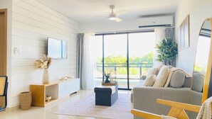 Ein modernes Wohnzimmer mit einer Couch, einem Fernseher, einem Deckenventilator und einem großen Fenster mit Blick auf Grünflächen.