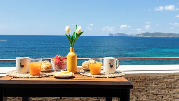 Ein Frühstückstisch mit Croissants, Orangensaft und einer Blumenvase, mit Blick auf die Küste.