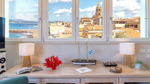 Ein Zimmer mit Blick auf eine Küstenstadt, ein Holztisch mit einem Schiffmodell, ein rotes Korallenstück und eine Schale Muscheln.