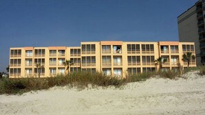 Et hotel med flere etager og fladt tag, beliggende på en sandstrand med sparsom vegetation.